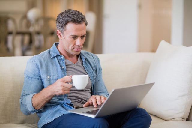 Man sitting on sofa in living room, using laptop while holding coffee cup. Ideal for illustrating remote work, home office setups, casual lifestyle, and technology use in everyday life.