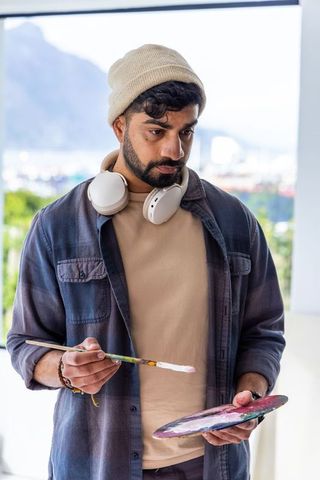 Male painter in studio preparing paint on palette and brush wearing beanie headphones