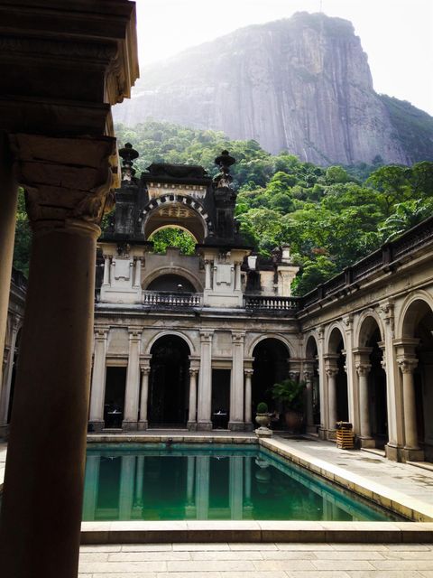 The photo showcases a serene historic courtyard with elegant European-style architecture and a reflective pool, set against a scenic mountain backdrop. The atmosphere is tranquil and timeless, making it perfect for use in travel magazines, architecture blogs, and tourism promotions. This image could also serve as an evocative background in presentations or as an inspirational scene in design projects.
