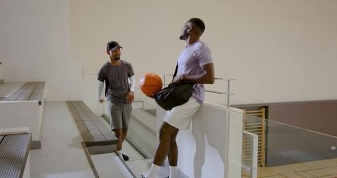 Diverse male friends chatting on gym bleachers holding basketball and duffel bag