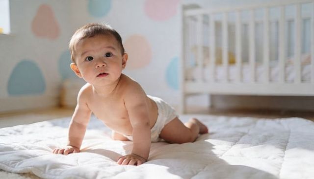 Curious baby crawling on quilted mat in sunlit nursery with white crib and pastel wall