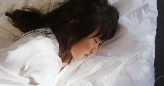 This serene scene of a young girl sleeping peacefully in white bedding can be used for promoting children's sleep products, bedtime stories, health and wellness articles, and family-related content. It conveys comfort, rest, and tranquility, making it ideal for use in advertising sleep aids or interior design for children's bedrooms.