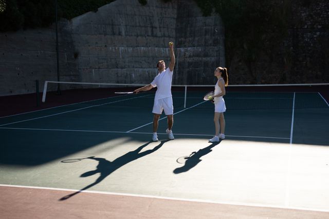 Caucasian man giving tennis instructions to a woman on a sunny outdoor court. Ideal for use in sports training materials, coaching advertisements, active lifestyle promotions, and summer sports campaigns.