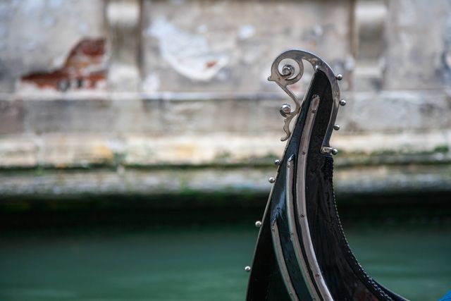 Detailed close-up of a Venetian gondola bow set against the backdrop of an old, rustic wall and tranquil water. Perfect for use in travel brochures, websites promoting Venice tourism, articles on Italian culture, or as a decorative image illustrating romantic or historic aspects of Venice.