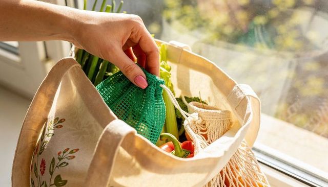 Woman packing reusable mesh produce bags into floral canvas tote on sunlit window sill