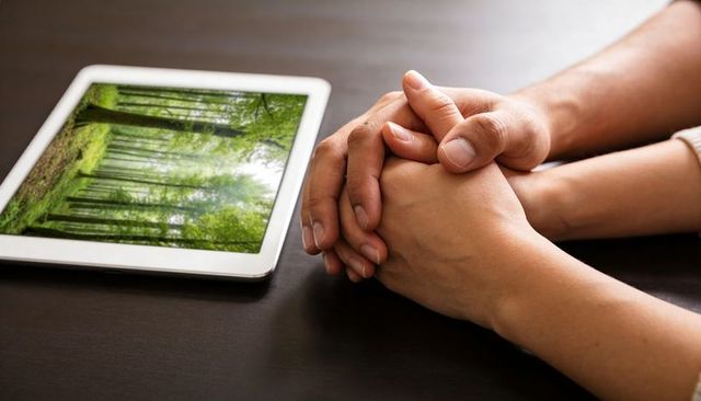 Couple clasping intertwined hands beside tablet displaying forest scene on dark wood table