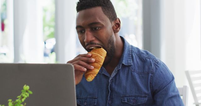 Young man eating croissant while working on laptop in cafe. Can be used for concepts of multitasking, casual dining, remote work, freelance lifestyle, and modern business practices.