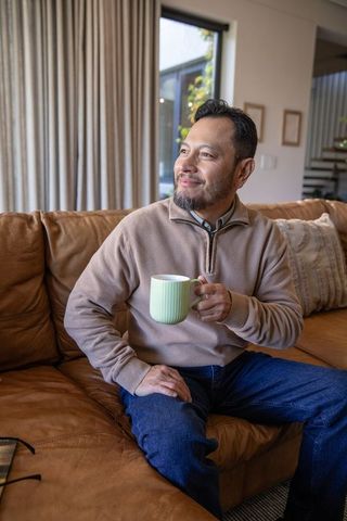 Mature asian man enjoying coffee on leather sofa, cozy morning home lifestyle portrait