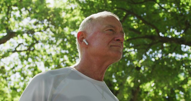 Elderly man engages in outdoor exercise under trees in daylight, wearing a white mesh shirt and wireless earbuds in a relaxed state. Ideal for fitness campaigns, senior health promotions, active lifestyle blogs, or advertisements for wireless technology.