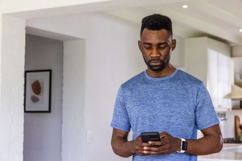 African man standing and using smartphone wearing white-band smartwatch in modern home