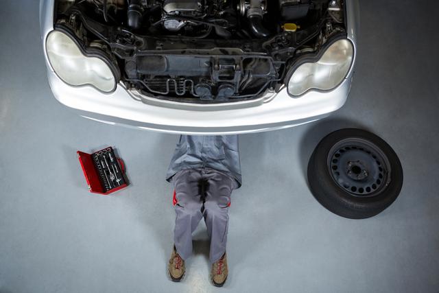 Female mechanic working underneath car, likely replacing or inspecting car parts. Scene includes a tire and a red toolbox nearby. Perfect for illustrating concepts of automotive repair, garage services, and women in skilled trades.