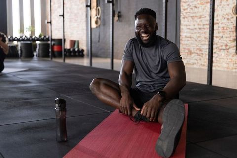 Laughing african american man stretching on red mat in gym with water bottle after workout