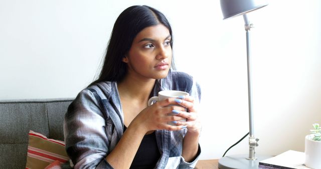 Young woman holding coffee cup in cozy living room and looking contemplative - Download Free Stock Images Pikwizard.com