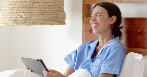 Young Woman in Blue Pajamas Laughing with Tablet in Cozy Bedroom