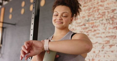 African american woman checking smartwatch while holding yoga mat in industrial gym