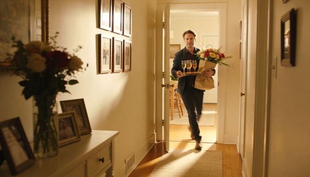 Man walking into home carrying bouquet and tray of champagne flutes, romantic arrival at doorway