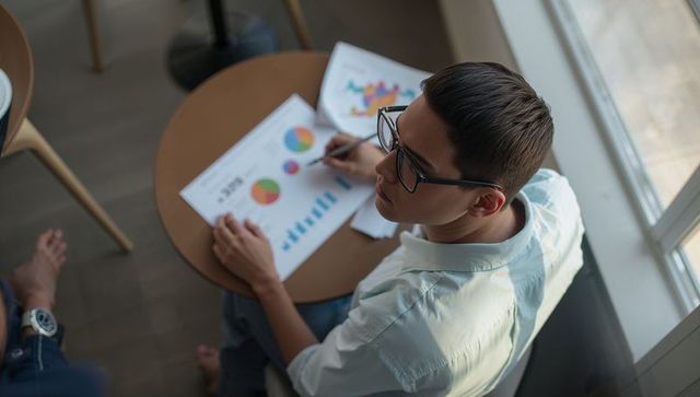 Asian student studying data charts at round cafe table wearing glasses and holding pen