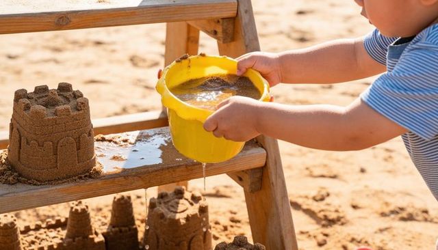 Child pouring water from yellow bucket over sandcastles on sunny beach with wooden ladder