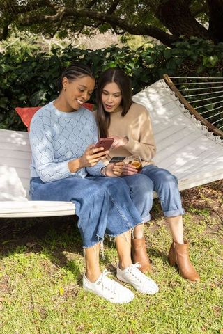 Diverse female friends sharing smartphone and credit card on backyard hammock, online shopping