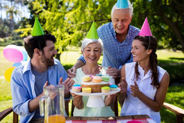 Family members of different generations celebrating a birthday outdoors in a park. They are wearing party hats and smiling while presenting a cake with colorful cupcakes. Ideal for use in advertisements, social media posts, and articles about family gatherings, celebrations, and outdoor activities.