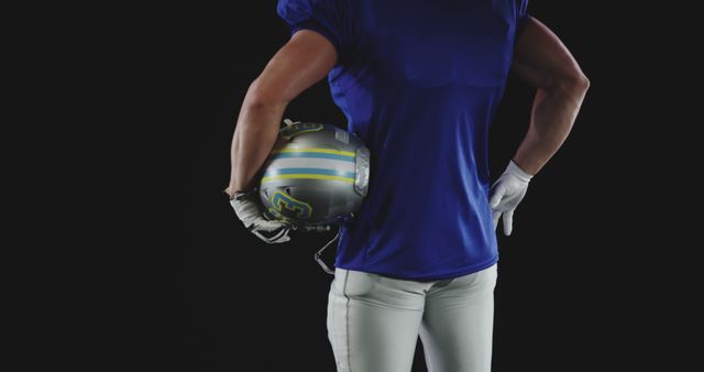 Football player stands with one hand holding helmet, showcasing strength and readiness. Ideal for sports blogs, fitness campaigns, ads promoting sports gear, or articles about perseverance in athletics.
