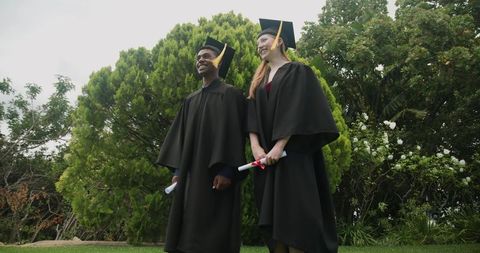 Diverse graduates wearing mortarboards and gowns holding ribboned diplomas on campus lawn