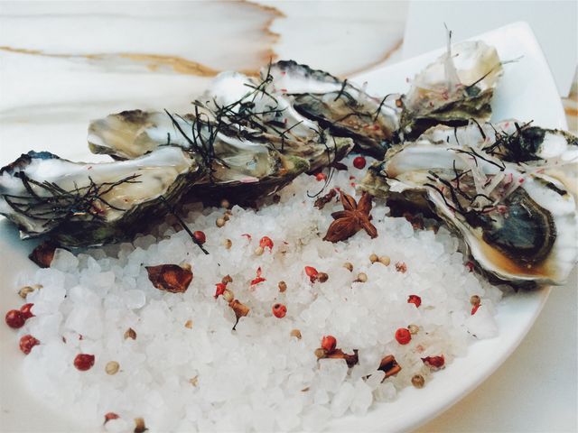 Fresh gourmet oysters displayed on plate, arranged on bed of seasoned rock salt with red and white peppercorns, and star anise. Ideal for use in culinary magazines, restaurant menus, food blogs, fine dining promotions and gourmet food marketing materials.