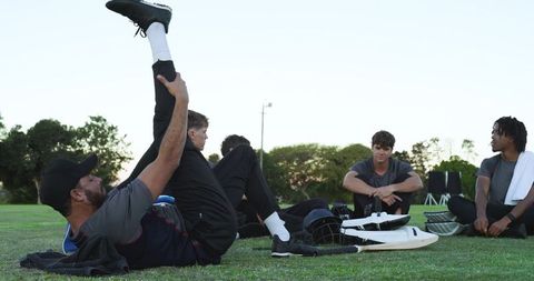 Cricket players stretching and recovering on grassy field at dusk with helmets and pads
