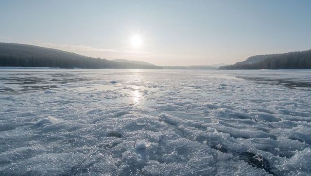 Icy lake surface glinting at sunrise with ice crystals and distant forested hills