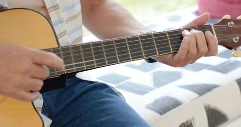 Mature man strumming acoustic guitar on sunlit couch, casual jeans and striped shirt
