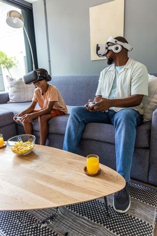 African american father and teenage son playing vr games on gray sofa, family bonding