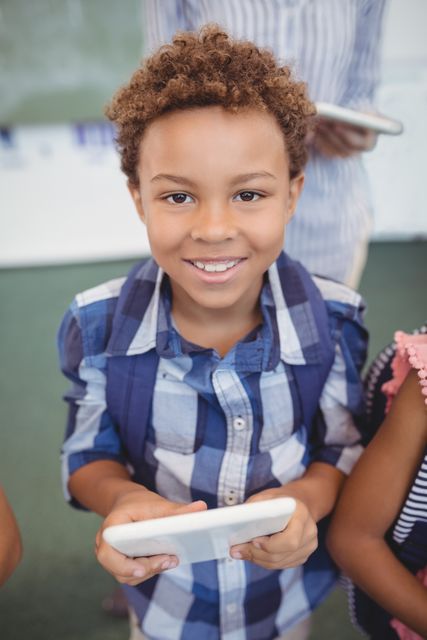 Young boy with curly hair holding a digital tablet and smiling in a classroom. Ideal for educational content, technology in education, school promotions, and interactive learning materials.