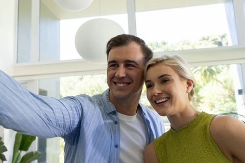 Young couple taking selfie in modern bright living room with large windows and greenery
