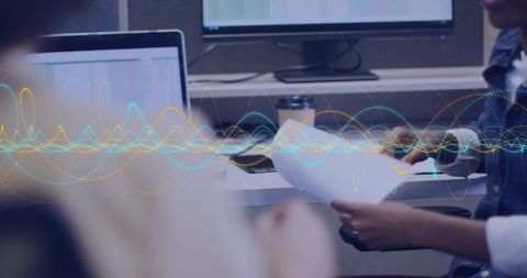 Woman reviewing documents at dual-monitor workstation with waveform data visualization overlay