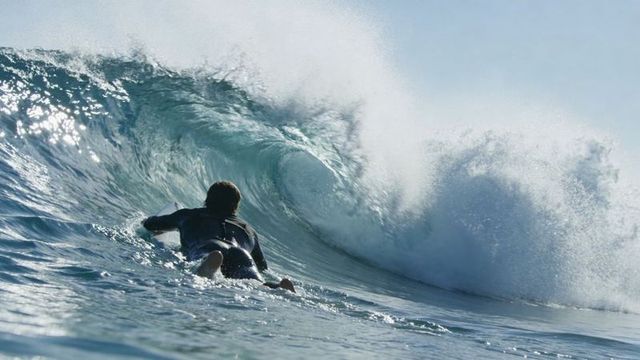 Paddling toward barrel surfer in wetsuit approaching tubular wave with sunlit spray