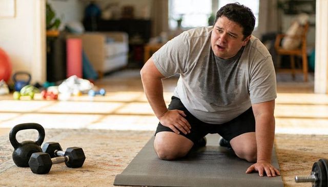 Overweight asian man panting after intense home workout on mat with kettlebell and weights