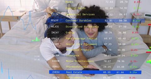 Young couple lying on bed sharing smartphone and laptop with stock market ticker overlay