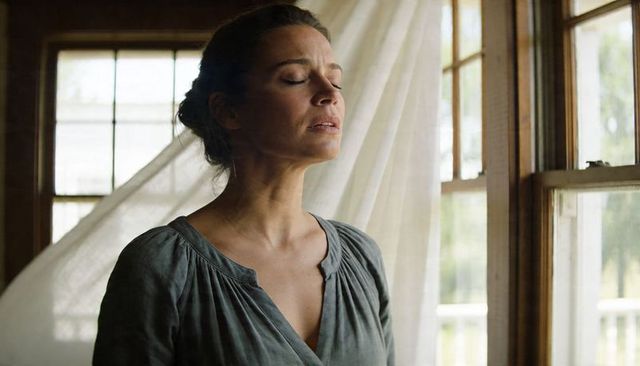 Serene woman standing by sunlit wooden window with billowing sheer curtains, eyes closed