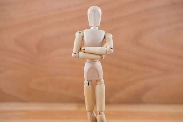 Wooden figurine standing with arms crossed against a neutral wooden background. Ideal for illustrating concepts of contemplation, creativity, and simplicity. Suitable for use in art class materials, creative projects, and minimalist design themes.