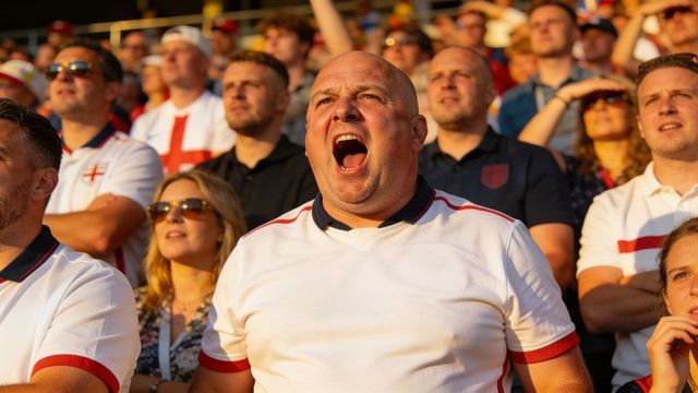 Bald fan shouting and cheering in sunlit stadium stands wearing england shirts with fellow fans