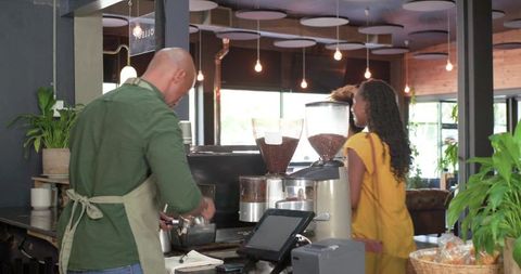 African barista preparing espresso and tamping portafilter for two women in cozy cafe