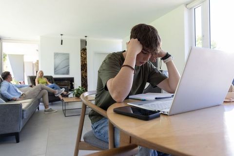 Teenager struggling with homework at dining table as parents relax in sunlit living room