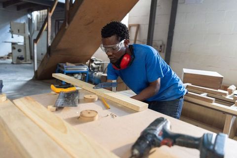 African american carpenter measuring clamping wooden plank wearing safety gear