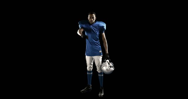 American football player in blue jersey and white pants holding helmet in one hand and pointing with other hand. Gloved and in full uniform on black background. Ideal for promoting sports events, team identity, athletic spirit, or equipment gear advertising.