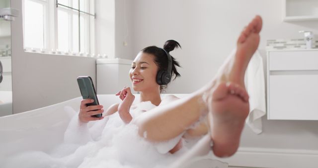 Image of portrait of smiling biracial woman with headphones and smartphone in bathtub in bubble bath. Health and beauty, leisure time, domestic life and lifestyle concept.