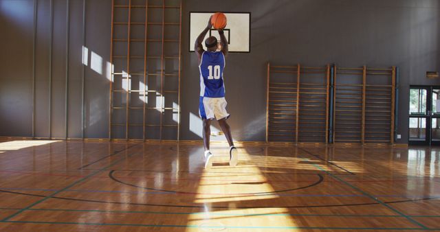 Young athlete in blue jersey shooting a basketball towards the hoop in an empty gymnasium with hardwood floors. Useful for content related to sports training, fitness, athletic practice, youth sports, or indoor activities. Ideal for articles, blogs, and advertisements focusing on basketball training, gymnasiums, or youth athletic programs.