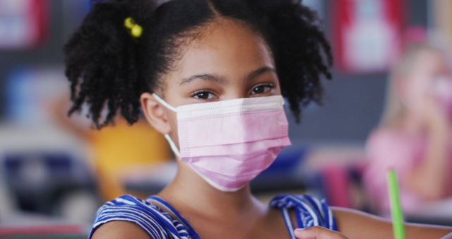 A young African American girl sits at her desk in a classroom, wearing a pink face mask and looking directly at the camera. This imagery can be used to depict themes of pandemic safety measures in schools, childhood education during COVID-19, public health awareness, and the importance of hygiene in classrooms. Useful for educational materials, public health campaigns, and news articles about pandemic impacts on students.