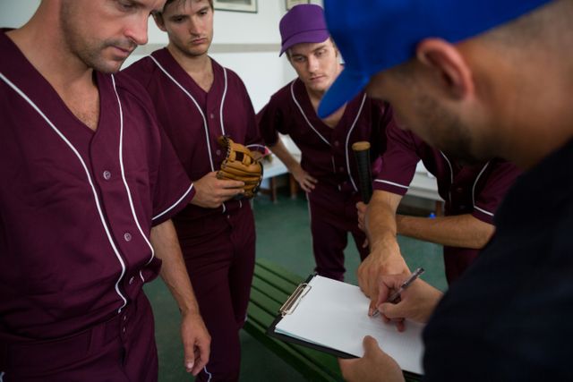 Baseball team players in maroon uniforms gather around their coach who is writing on a clipboard. This scene captures the moment of strategic planning and teamwork before a game. Ideal for use in sports-related articles, teamwork and leadership concepts, and promotional materials for baseball events.