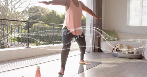 Senior woman balancing in warrior yoga pose on sunlit balcony mat with dog resting nearby