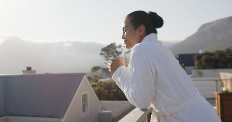 Asian woman standing on balcony holding mug, gazing over rooftops to mountain sunrise